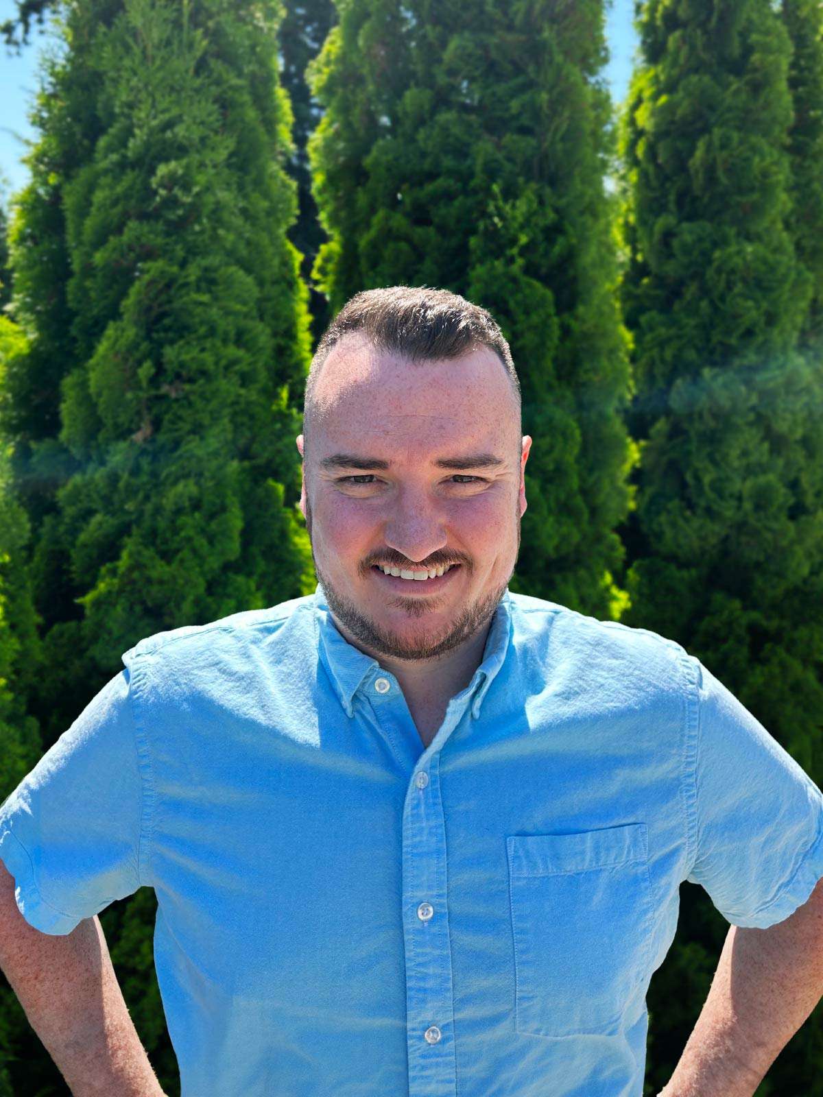 A man in a light blue, short-sleeve button-up shirt stands outdoors in front of tall, green trees, smiling at the camera on a sunny day.