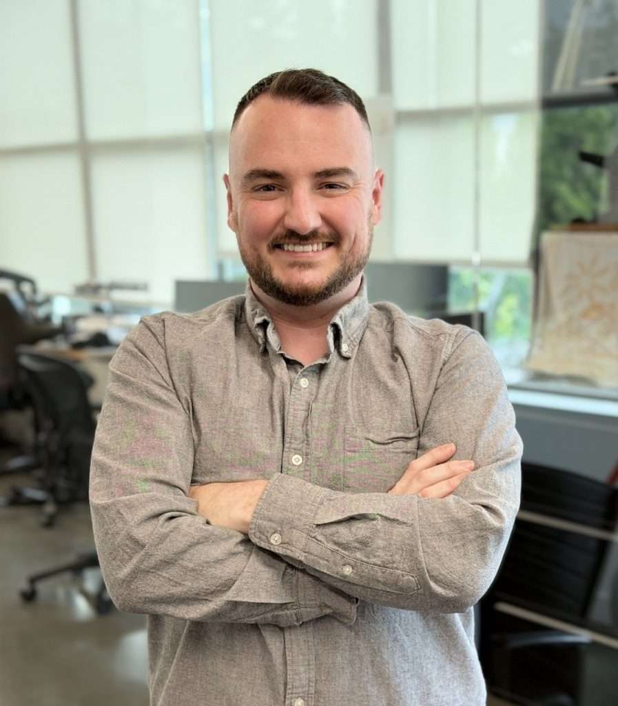 A man with short hair and a trimmed beard stands indoors with arms crossed, smiling. He wears a gray button-up shirt. Office furniture and large windows with blinds are visible in the background.