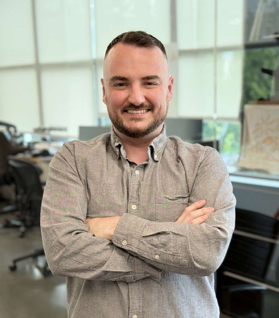 A man with short hair and a trimmed beard stands indoors with arms crossed, smiling. He wears a gray button-up shirt. Office furniture and large windows with blinds are visible in the background.