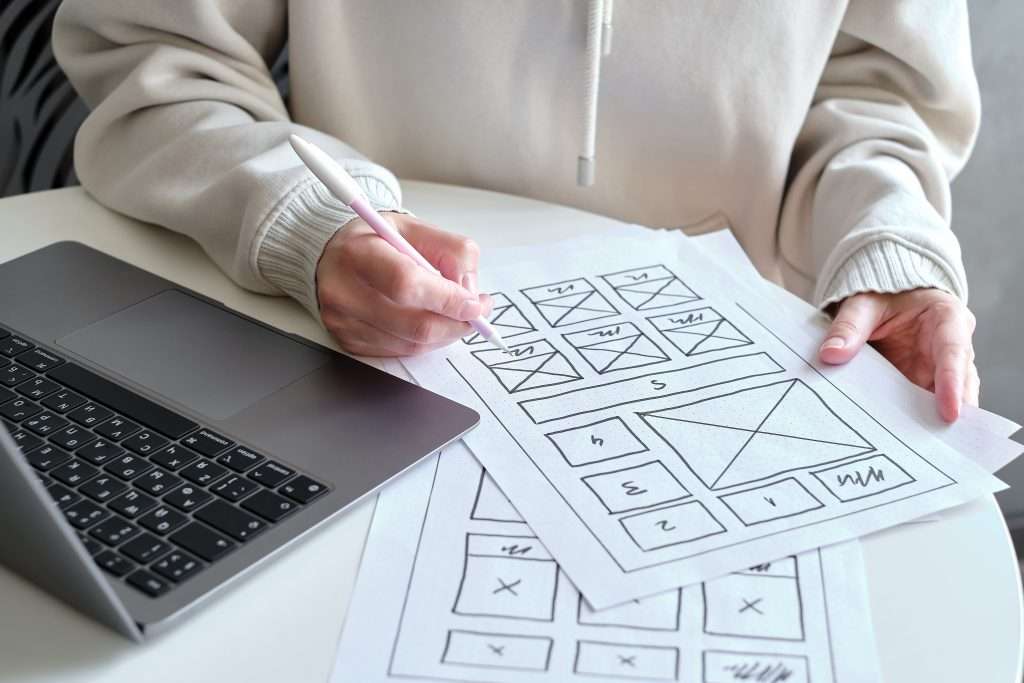 A person designs website layouts with a pen on printed wireframe sketches, sitting beside an open laptop on a white desk.