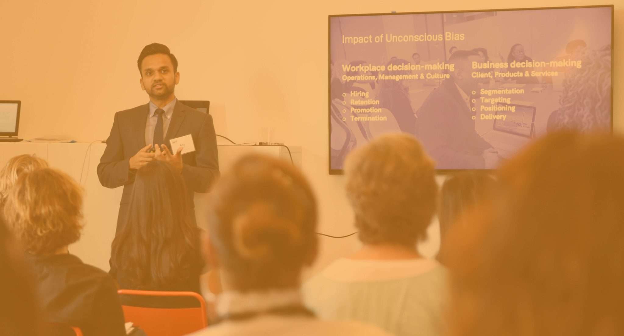 A man stands and presents to an audience in a classroom, with a slide titled Impact of Unconscious Bias displayed on a screen behind him. The slide lists effects on workplace and business decision-making.