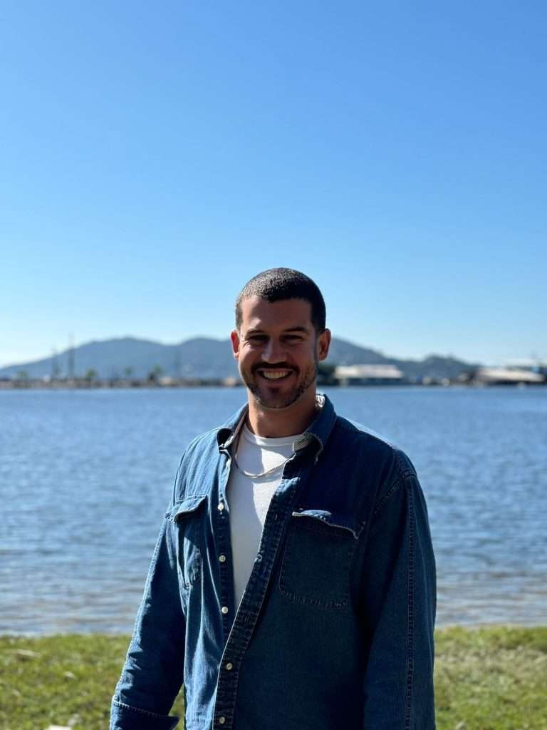 A man in a blue denim shirt stands smiling by a calm body of water, with grassy shoreline in the foreground and distant hills under a clear blue sky in the background.