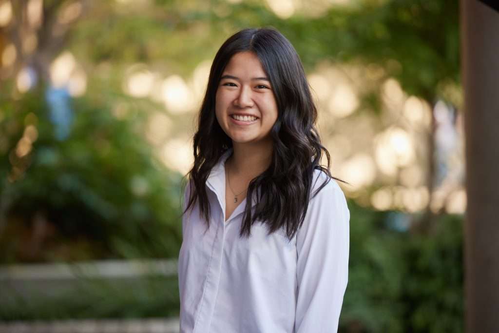 A young woman with long, wavy black hair smiles while standing outdoors. She is wearing a white button-up shirt, with green foliage and sunlight softly blurred in the background.