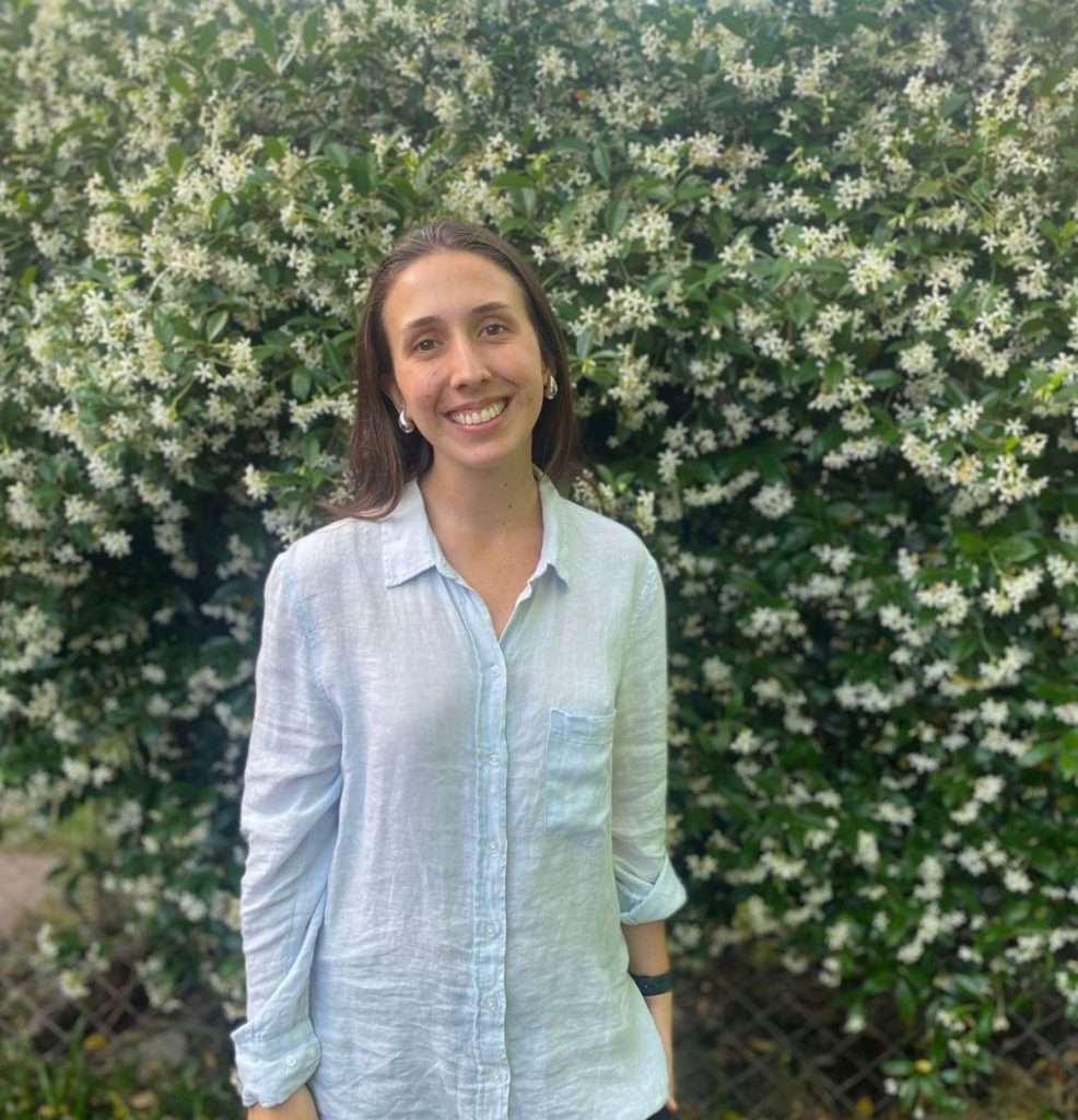 A woman with long brown hair, wearing a light blue button-up shirt, stands smiling in front of a lush green bush covered with many small white flowers.