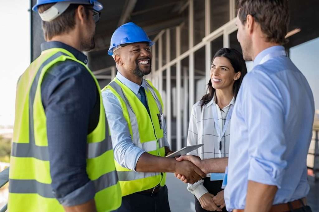 Two men in safety vests and hard hats shake hands with two professionals, one woman and one man, on a construction site. All are smiling, suggesting a successful meeting about digital marketing for contractors.