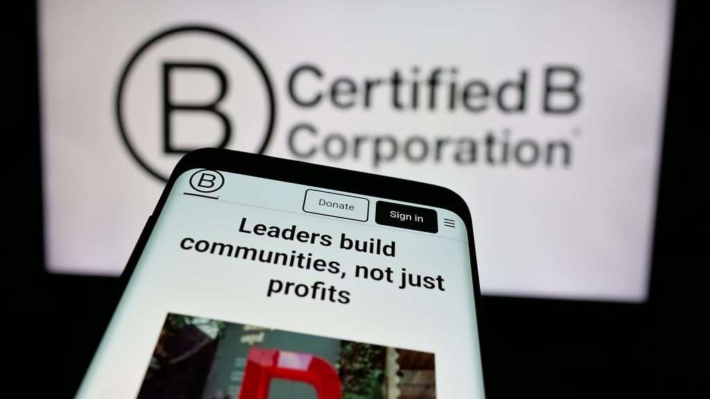 A smartphone screen displays a website with the headline Leaders build communities, not just profits and buttons for Donate and Sign in. In the background is a blurred Certified B Corporation logo.