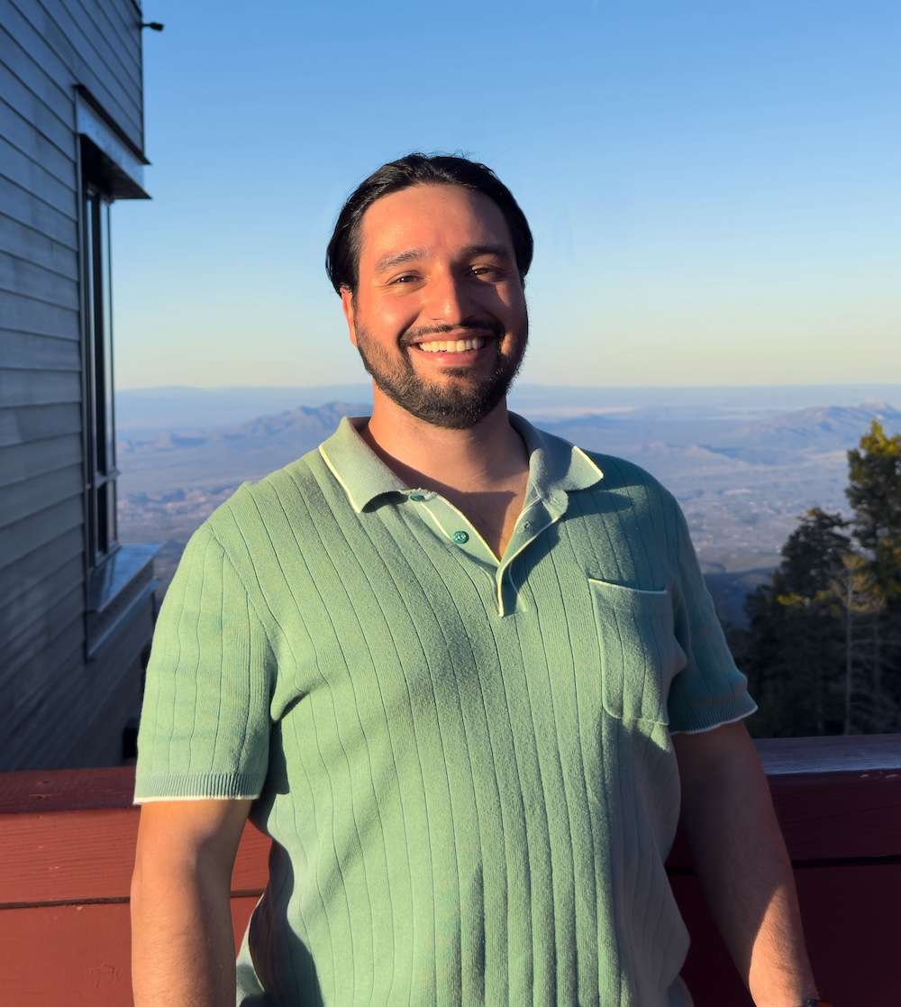 A smiling man with dark hair and a beard wears a light green short-sleeved polo shirt, standing outside on a balcony with mountains and a clear sky in the background. Sunlight casts a warm glow on him.