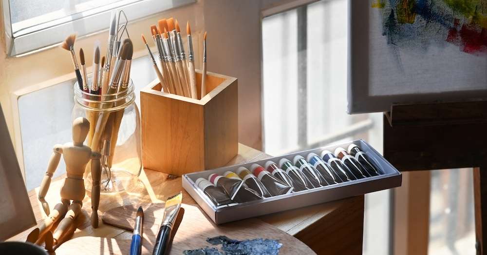 Paintbrushes in jars, tubes of paint, a wooden artist mannequin, and part of a canvas with colorful paint on a sunlit table create a cozy and creative art studio—perfect for brainstorming solutions to questions like why can't I find my business online.