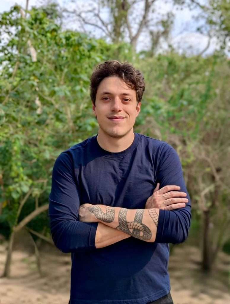 Carlos Bermudez Headshot - A person with short brown hair, wearing a navy blue long-sleeve shirt, stands outdoors with arms crossed, smiling. They have visible tattoos on their forearms. Green trees and foliage fill the background.