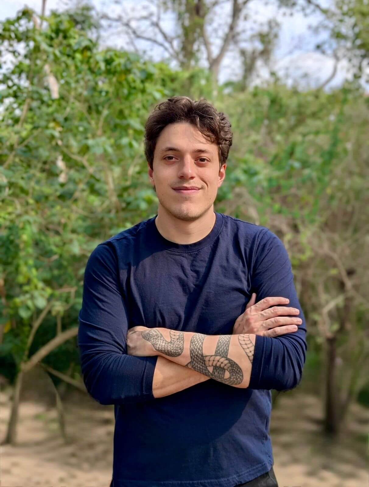 Carlos Bermudez Headshot - A person with short brown hair, wearing a navy blue long-sleeve shirt, stands outdoors with arms crossed, smiling. They have visible tattoos on their forearms. Green trees and foliage fill the background.