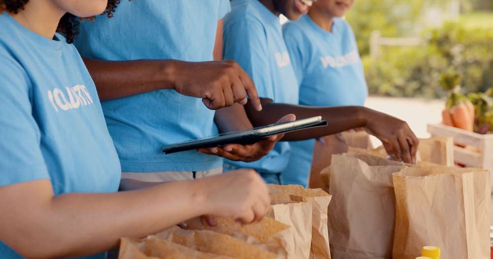 Four people in matching blue shirts pack brown paper bags with food items outdoors; one person is holding and pointing at a tablet while others fill bags, suggesting a volunteer or charity food distribution event.
