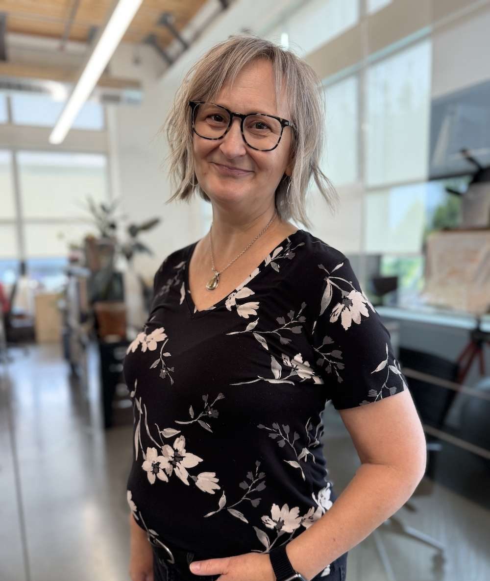 A smiling woman with short, wavy gray hair and glasses stands indoors, wearing a black floral shirt and black pants. The background shows a bright, modern office space with large windows and plants.