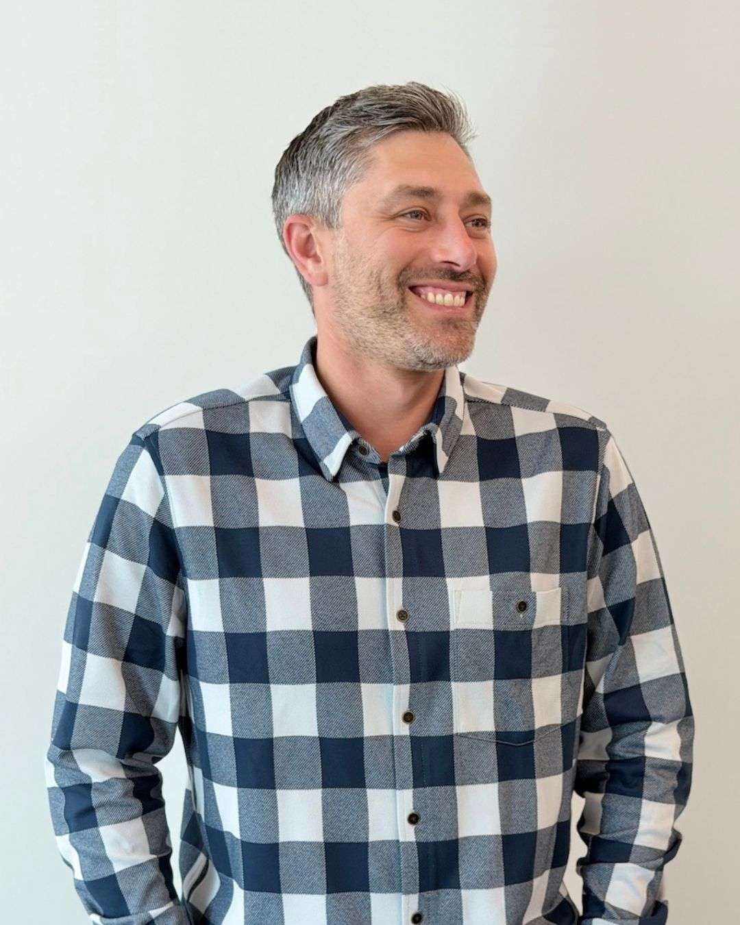 A man with short gray hair and a beard, wearing a blue and white checkered shirt, smiles while looking to the side against a plain light background.