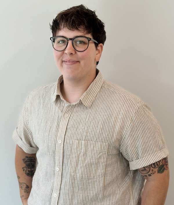 Person with short dark hair, glasses, and a beige striped shirt stands smiling with hands in pockets. Their arms have visible tattoos and they wear a smartwatch. The background is plain and light-colored.