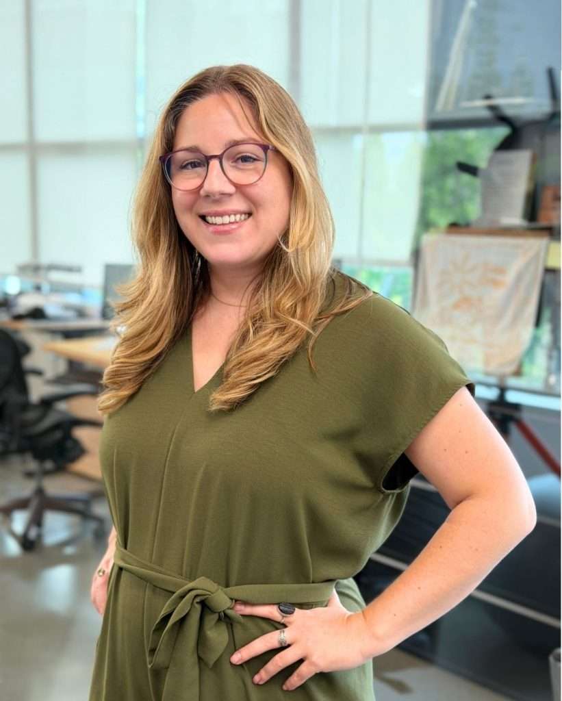 A woman with long, light brown hair and glasses smiles, standing indoors in an office wearing a green dress with a tie at the waist. Desks, chairs, and bright windows are visible in the background.